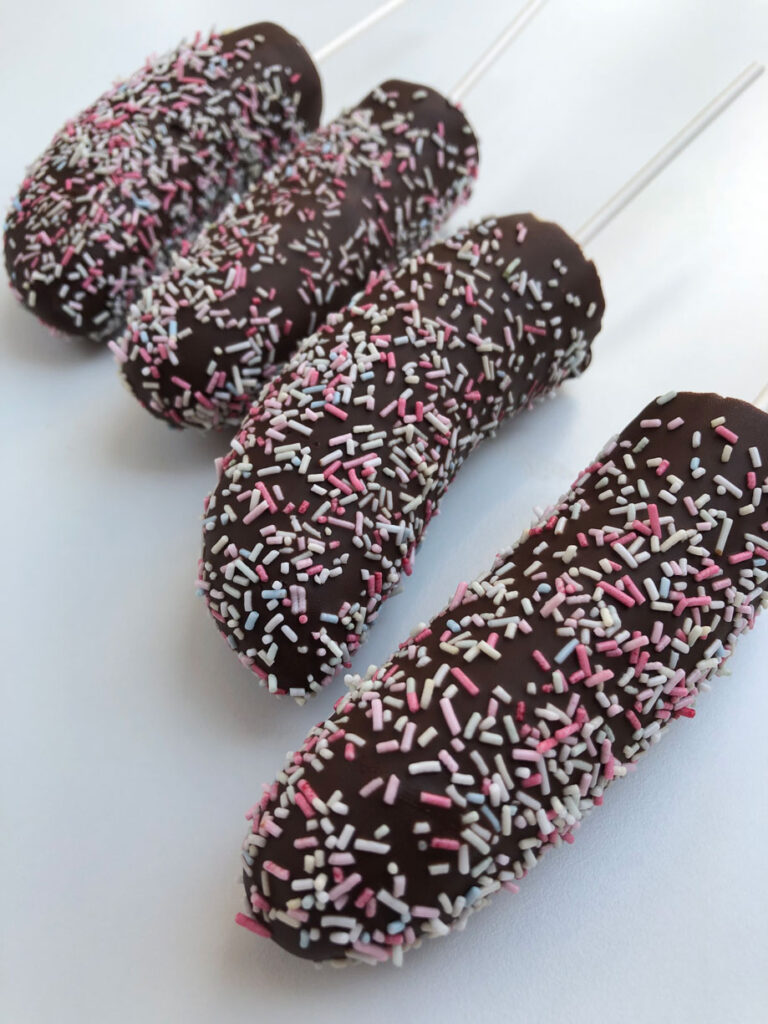 Four frozen chocolate-covered bananas decorated with sprinkles on a white surface.
