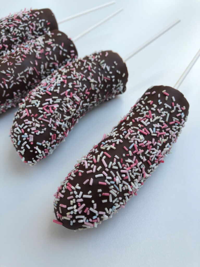 Four frozen chocolate-covered bananas decorated with sprinkles on a white surface.