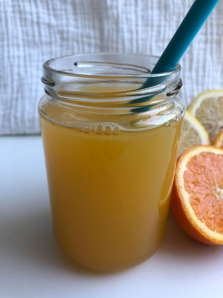 Homemade orange lemonade in a glass with a straw, orange and lemon halves beside the glass