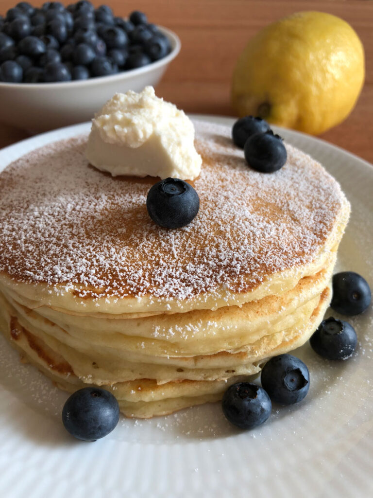 Lemon ricotta pancakes on a plate served with powdered sugar, ricotta, and fresh blueberries, in the background a lemon and a bowl of fresh blueberries.