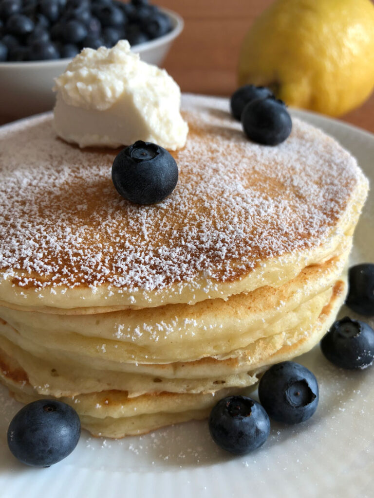 Lemon ricotta pancakes on a plate served with powdered sugar, ricotta, and fresh blueberries, in the background a lemon and a bowl of fresh blueberries.