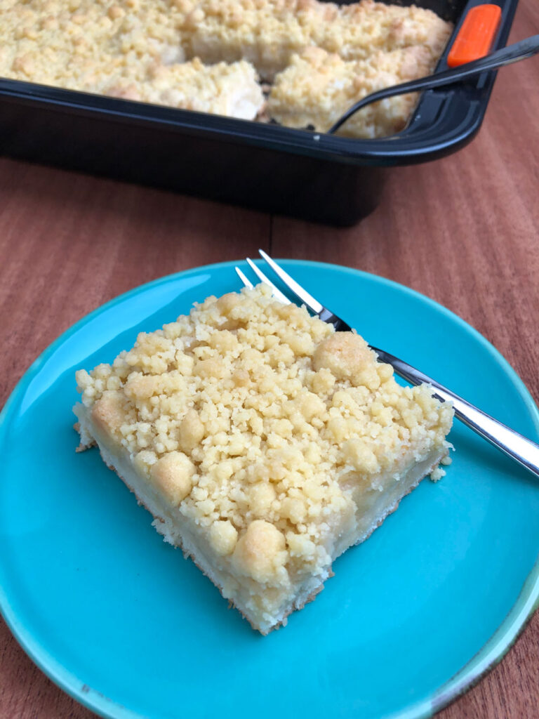 A piece of German apple streusel sheet cake sitting next to a fork on a plate, with the sliced cake in the baking pan in the background.
