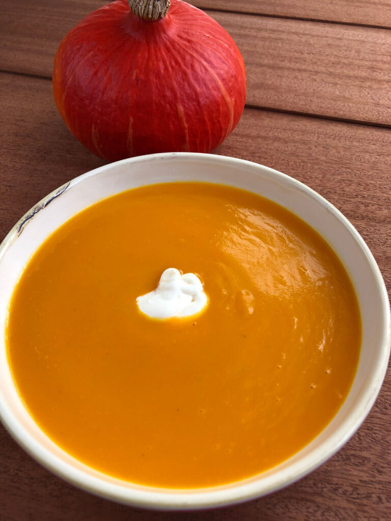 Easy pumpkin soup served in a bowl with a dollop of crème fraîche in the center, and a pumpkin in the background.