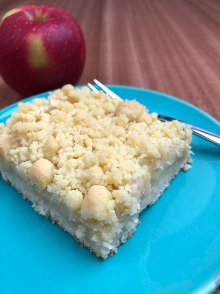 A piece of German apple streusel sheet cake sitting next to a fork on a plate, with a whole red apple in the background.
