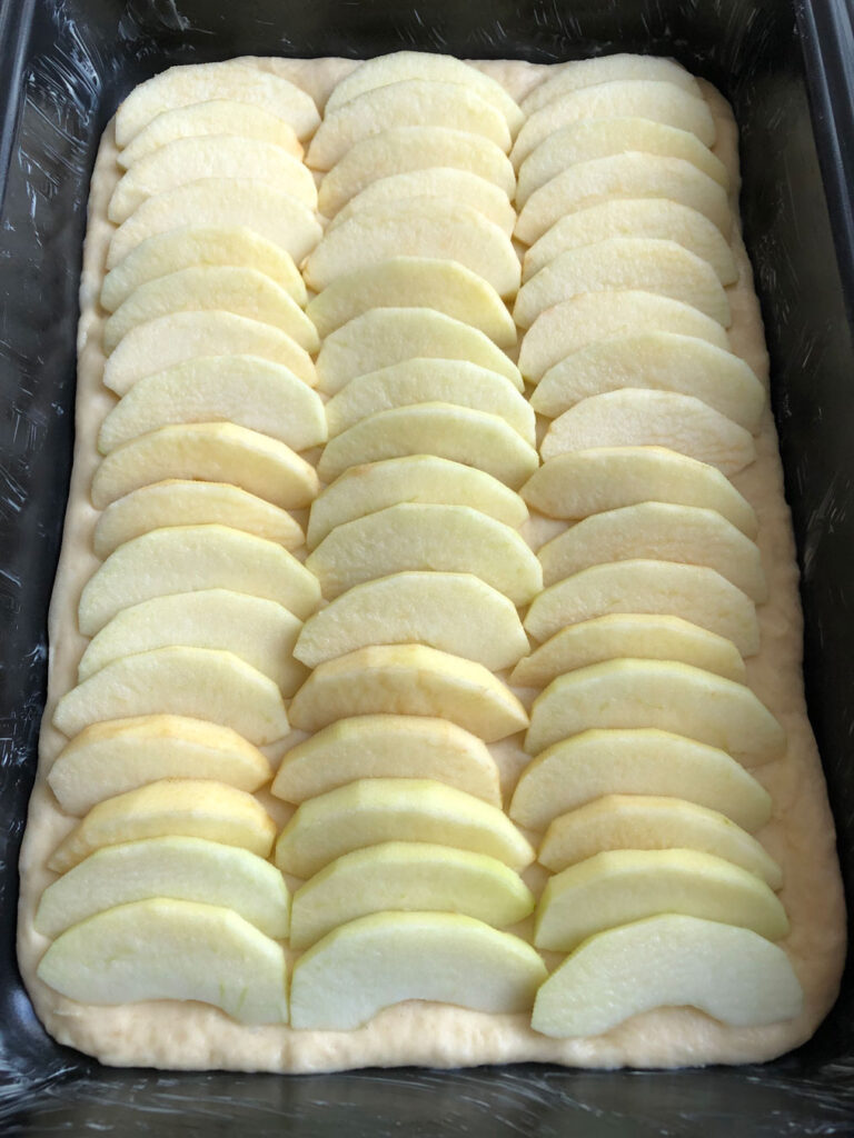 Yeast dough base for a German apple streusel sheet cake, topped with sliced apples, before being sprinkled with streusel.