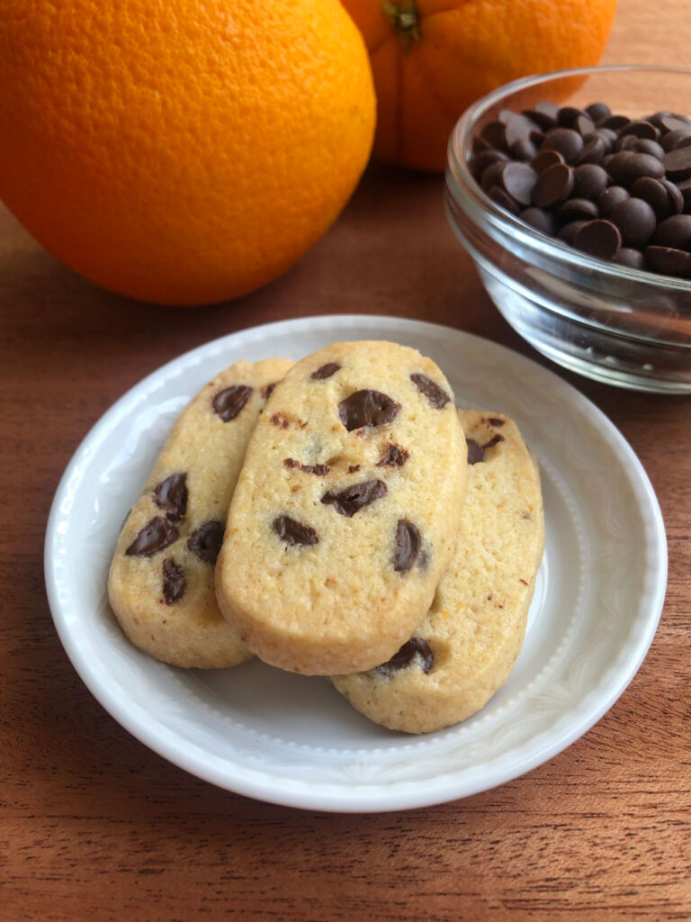 Chocolate chip orange slice and bake cookies on a plate, chocolate chips in a bowl and oranges in the background.