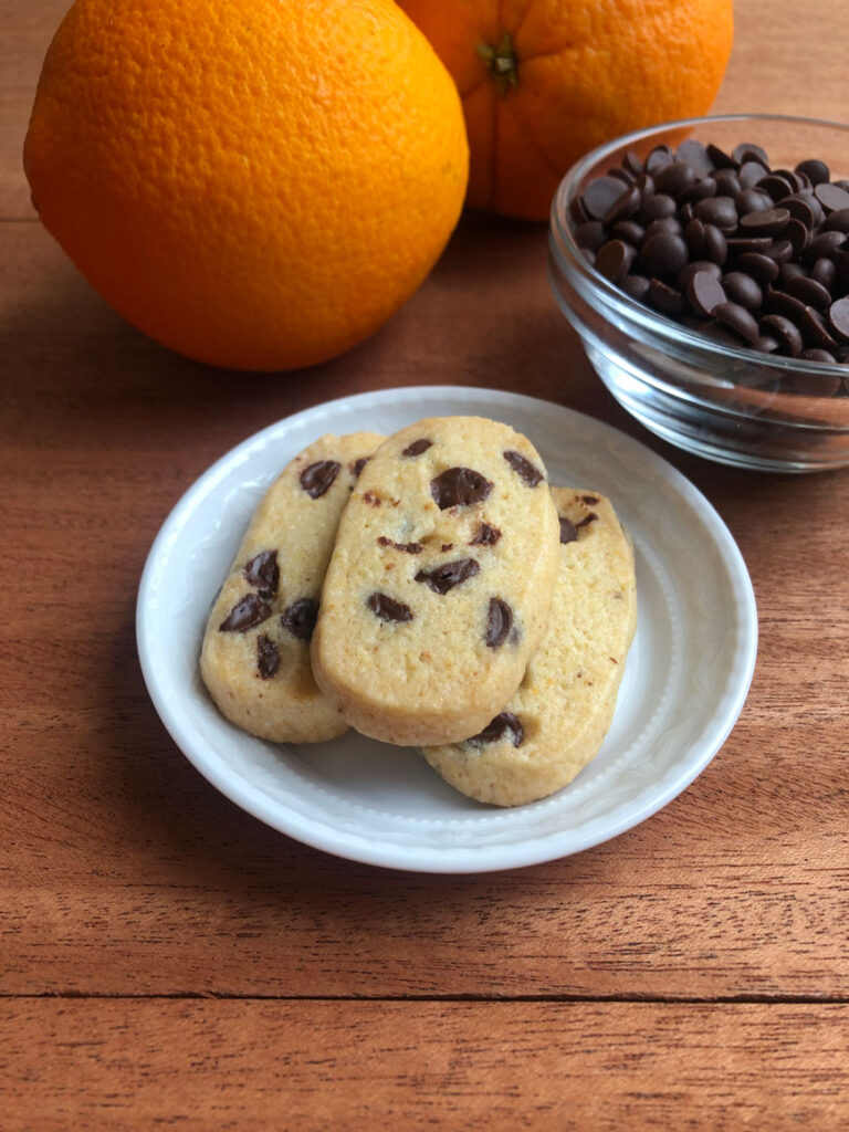 Chocolate chip orange slice and bake cookies on a plate, oranges and chocolate chips in a bowl in the background.