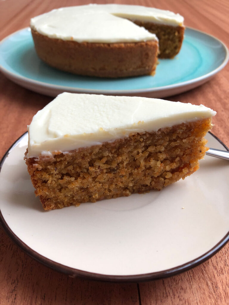 A slice of carrot cake with cream cheese frosting on a plate, with a carrot cake on a cake plate in the background.
