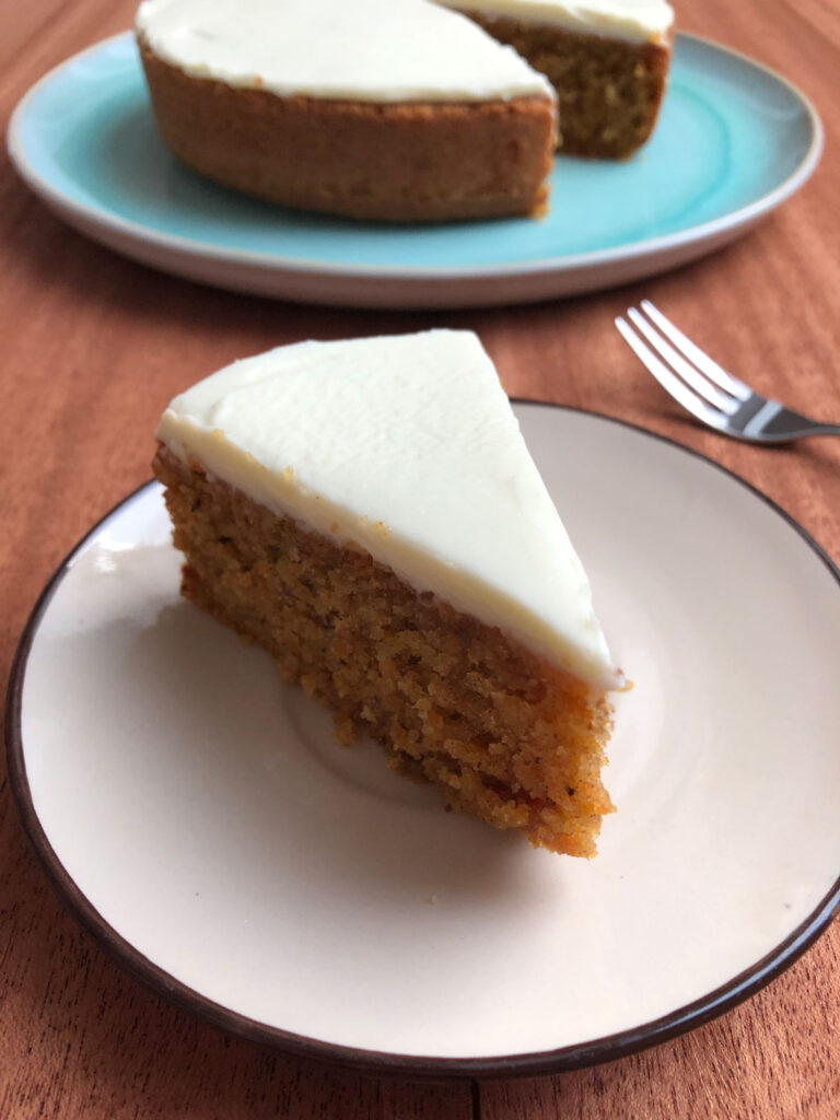 A slice of carrot cake with cream cheese frosting on a plate, with a carrot cake on a cake plate in the background.