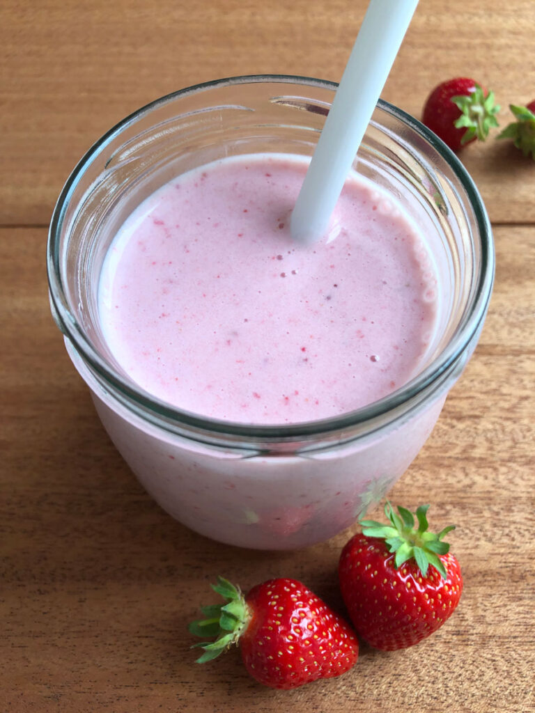 Creamy strawberry smoothie in a mason jar with a straw in it, fresh strawberries in the background.
