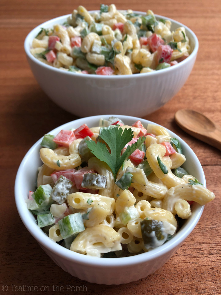 Classic Macaroni Salad Recipe Colorful classic macaroni salad in a small bowl, decorated with a flat-leaf parsley leaf, a large serving bowl with more macaroni salad in the background.