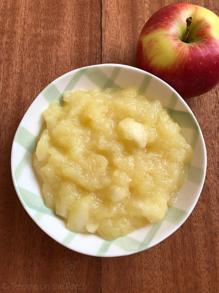 Bird's-eye view of easy apple compote in a green and white checkered bowl, with a fresh looking red apple in the background.