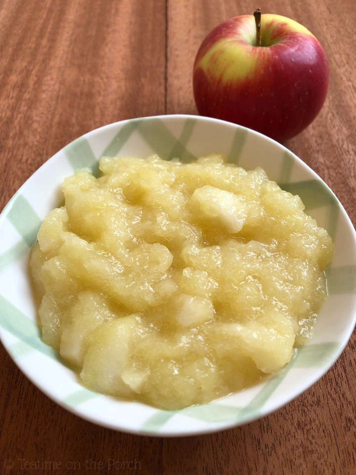 Apple compote in a green and white checkered bowl, with a fresh looking red apple in the background.